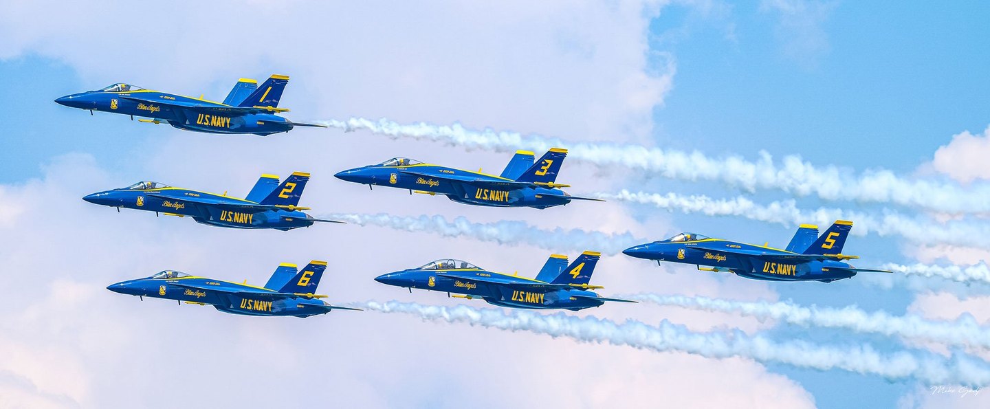 Six blue and yellow U.S. Navy Blue Angels jets fly leftward in formation, trailing white smoke across a blue sky filled with pale clouds.