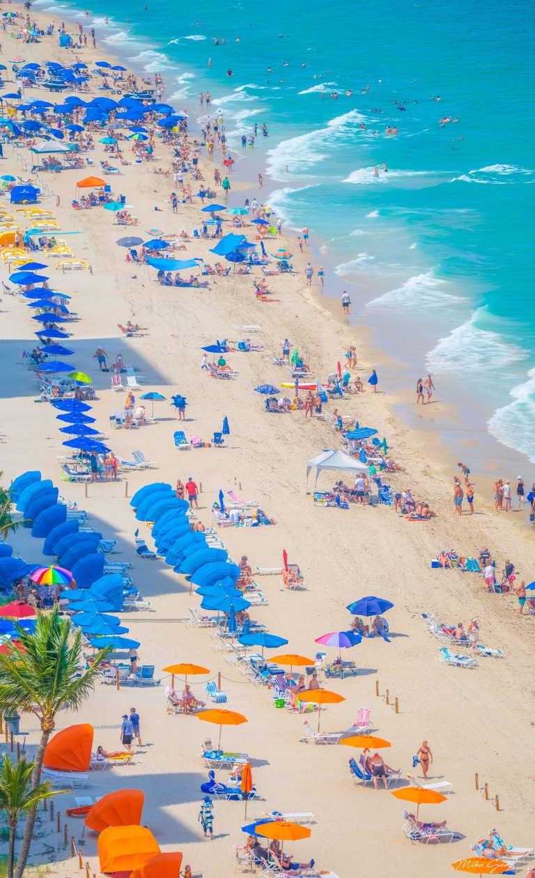 An elevated view of a crowded sandy beach shows blue and orange umbrellas, lounge chairs, palm trees, and swimmers in turquoise surf.
