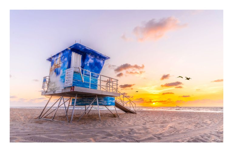 A blue lifeguard tower painted with clouds stands on an empty sandy beach as birds fly above the ocean and a golden sun rests near the horizon.
