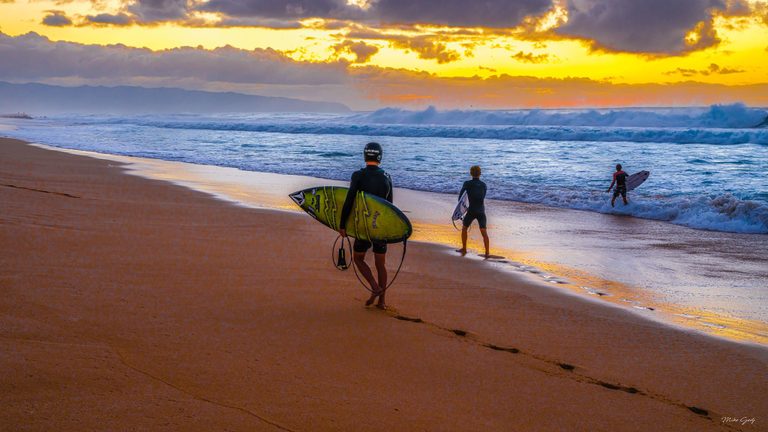 Three surfers carry boards toward breaking waves on a sandy beach beneath a golden, cloud-filled sky, with distant mountains along the horizon.
