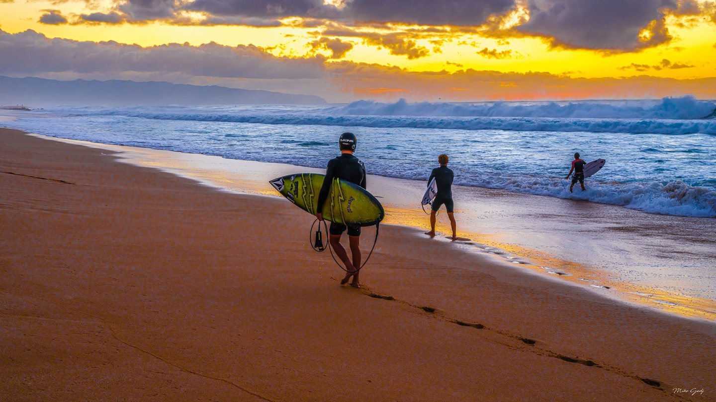 Three surfers carry boards toward breaking waves on a sandy beach beneath a golden, cloud-filled sky, with distant mountains along the horizon.