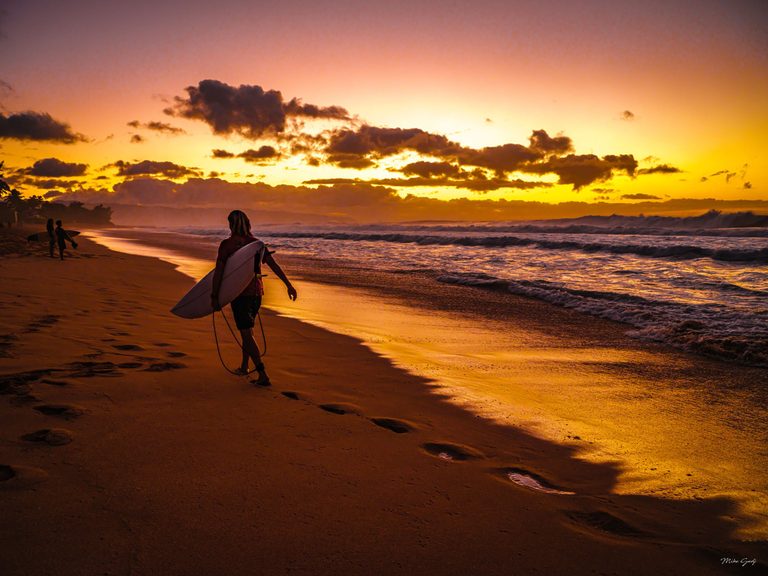 A surfer carries a surfboard along a sandy beach beneath an orange and gold sky, with breaking waves to the right and two figures farther along the shore.