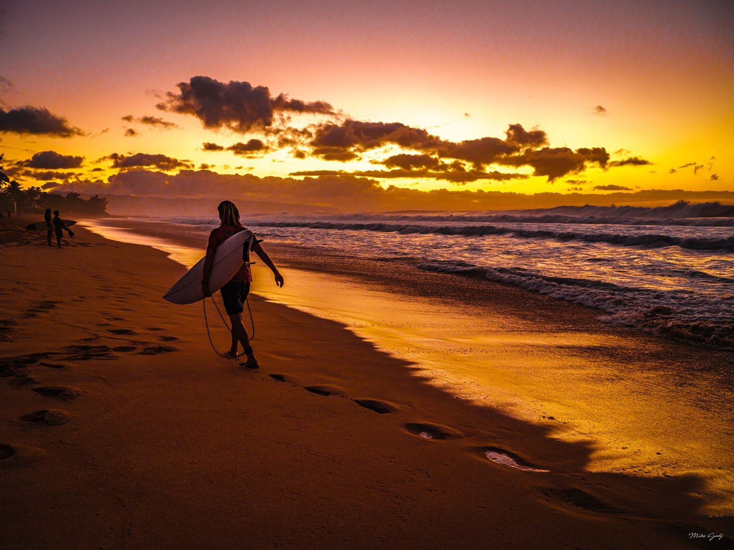 A surfer carries a surfboard along a sandy beach beneath an orange and gold sky, with breaking waves to the right and two figures farther along the shore.