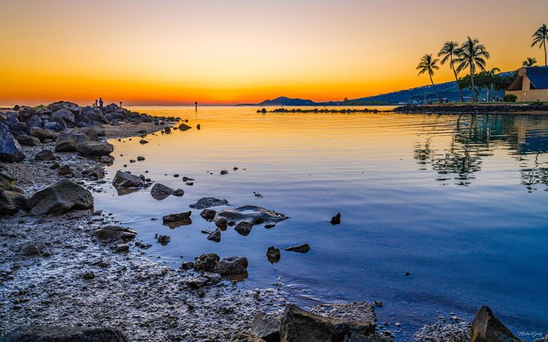 A rocky shoreline curves around calm blue water reflecting an orange sunset, with palm trees, a waterfront building, and distant hills across the bay.