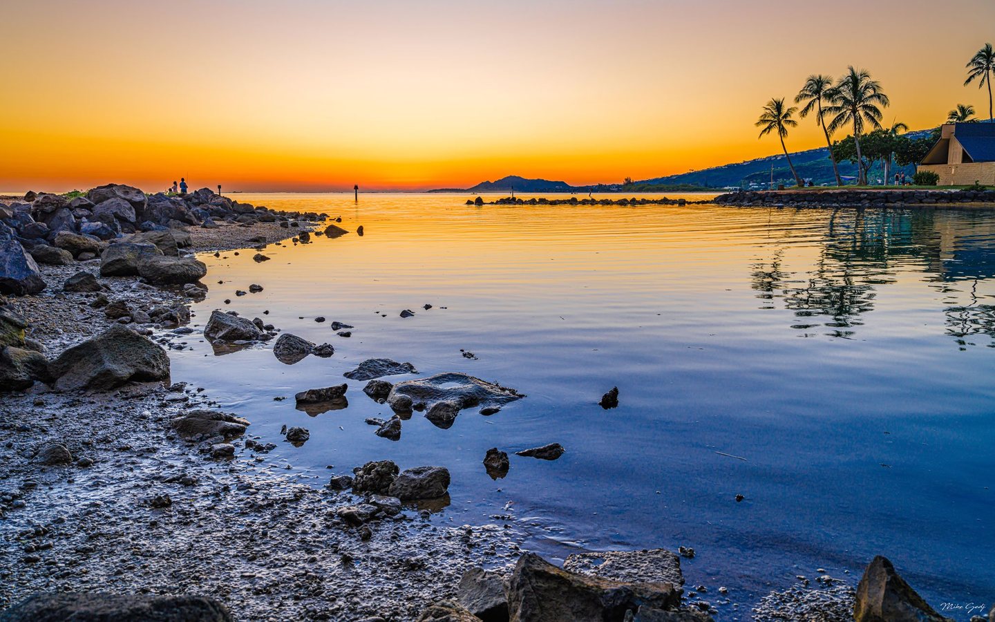 A rocky shoreline curves around calm blue water reflecting an orange sunset, with palm trees, a waterfront building, and distant hills across the bay.
