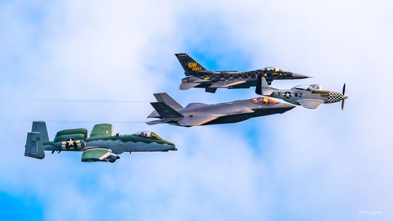 An A-10, F-35, F-16, and P-51 Mustang fly toward the right in a staggered formation against a pale blue, cloud-filled sky.