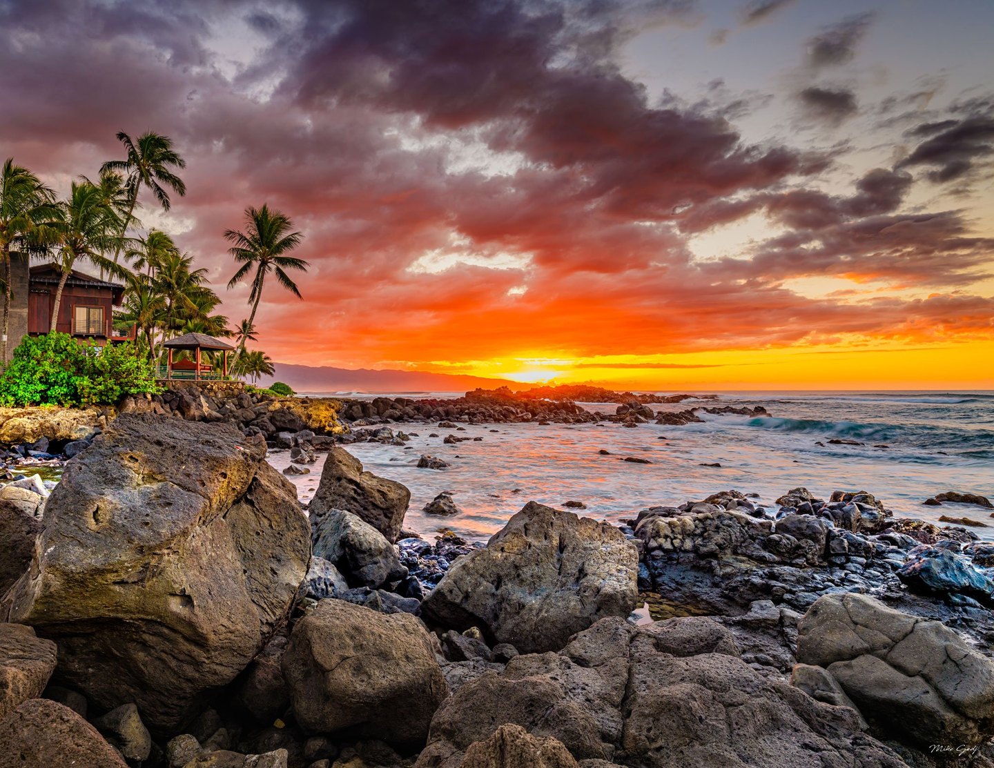 Orange and purple sunset clouds stretch over a rocky tropical shoreline, with palm trees and waterfront buildings on the left and waves breaking offshore.