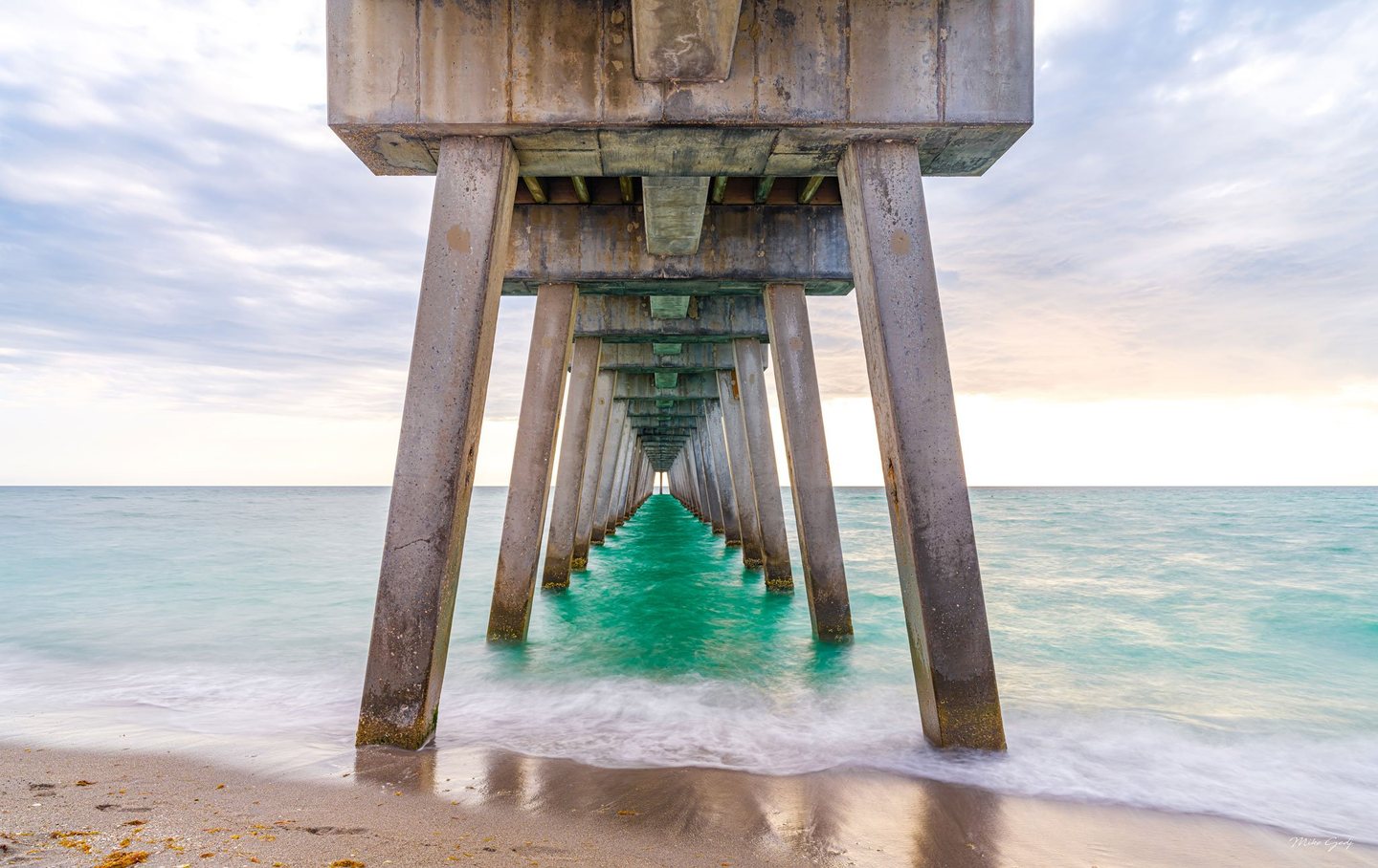 A view beneath a concrete pier shows repeating pillars receding over turquoise seawater, with soft surf washing onto wet sand beneath a pale, cloudy sky.