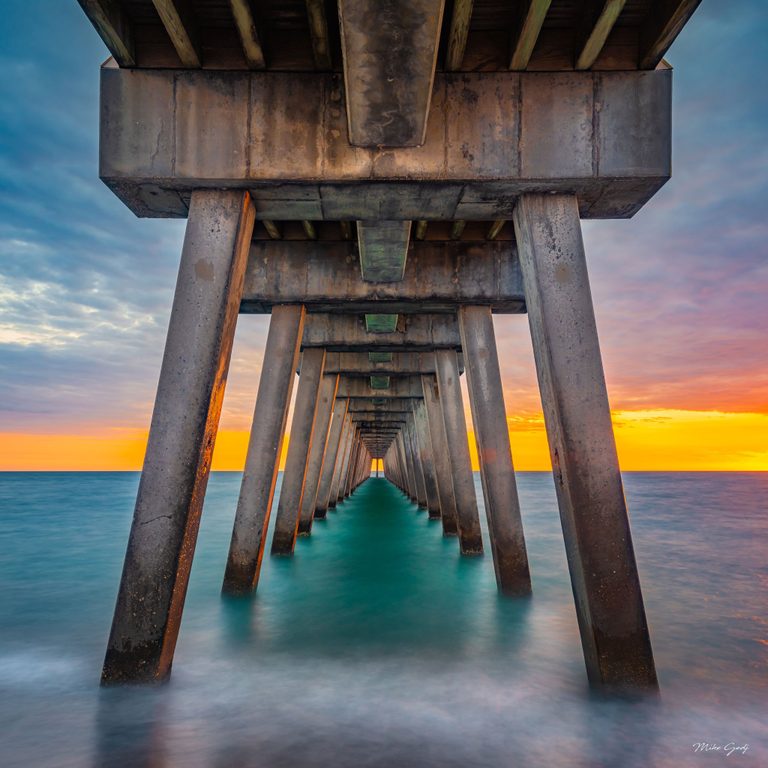 Rows of weathered concrete pillars beneath a pier converge toward the horizon above turquoise ocean water under an orange and blue sky.