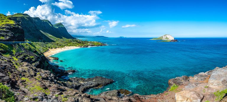 Turquoise ocean water curves around a sandy beach beneath steep green cliffs, with dark foreground rocks and a small offshore island under a bright blue sky.