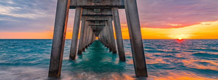 Concrete pier supports recede toward the horizon above turquoise waves, with a low sun and orange-pink clouds to the right.