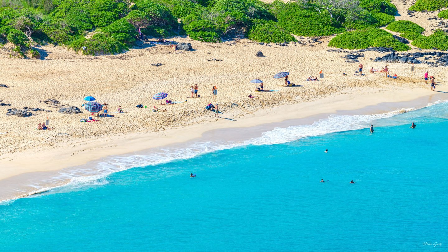 An elevated view shows swimmers in turquoise water beside a sandy beach dotted with striped umbrellas, sunbathers, dark rocks, and dense green vegetation.