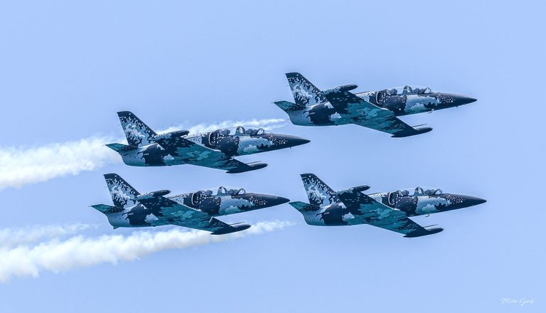Four jets with blue, black, and white patterned paint fly in close formation toward the right against a pale blue sky, with white smoke trailing behind.