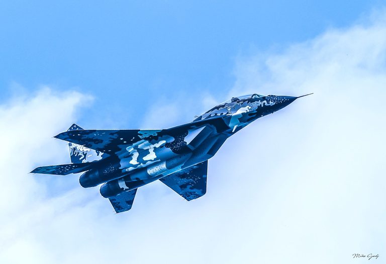 A blue-and-white patterned twin-engine fighter jet banks overhead against a blue sky with soft white clouds.
