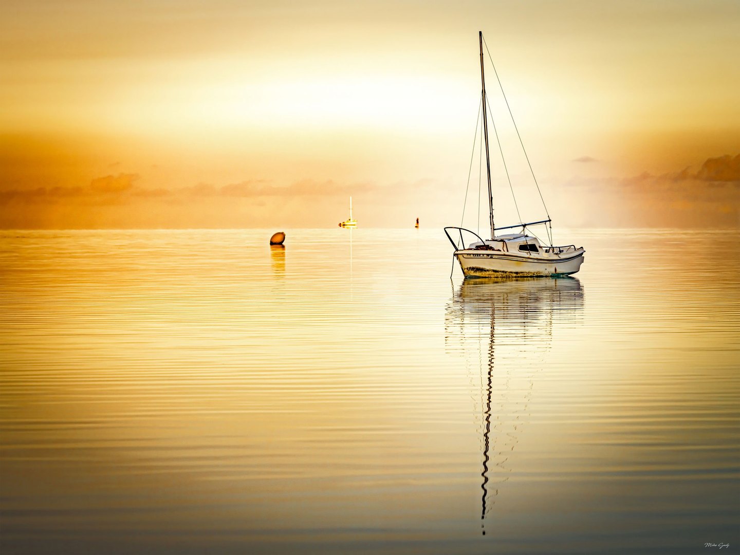 A white sailboat rests on calm golden water, its tall mast reflected in gentle ripples, with a buoy and another boat in the distance.