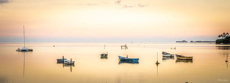 Small moored boats and a sailboat reflect in calm golden water beneath a peach sky, with distant rocky islets and a palm-lined shore to the right.