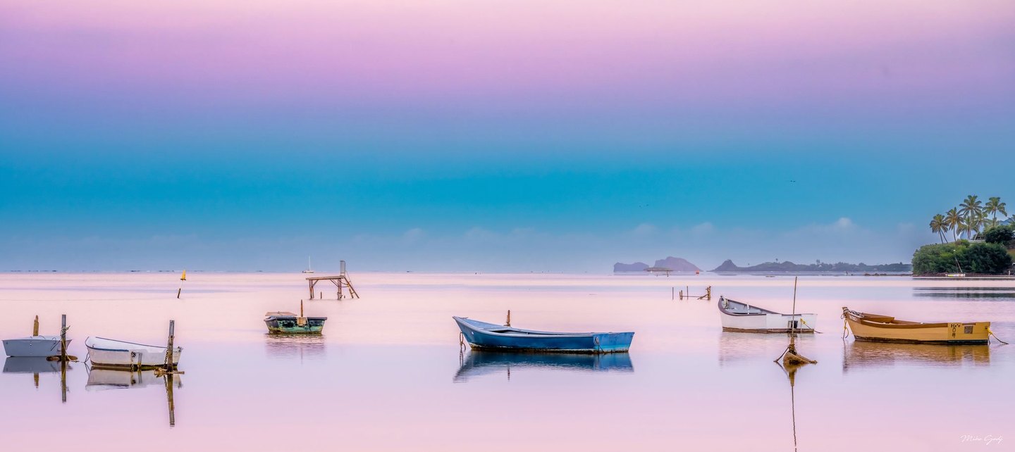 Small blue, white, green, and yellow boats lie moored on calm pink-toned water beneath a blue and lavender sky, with distant islands and palms along the right shore.