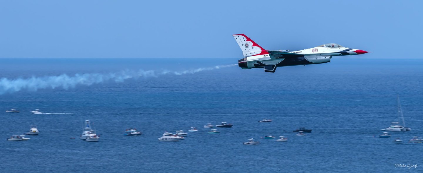 A red, white, and blue U.S. Air Force Thunderbirds jet flies above an ocean dotted with boats, trailing white smoke beneath a clear sky.