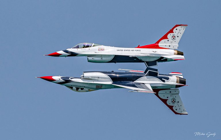 Two red, white, and blue U.S. Air Force Thunderbirds jets fly in close formation against a clear blue sky, with the lower aircraft inverted.