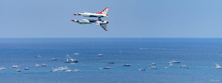 Two red, white, and blue jets fly in close formation, one inverted beneath the other, above a blue ocean dotted with motorboats.