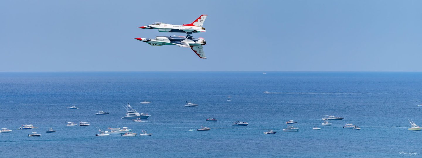 Two red, white, and blue jets fly in close formation, one inverted beneath the other, above a blue ocean dotted with motorboats.