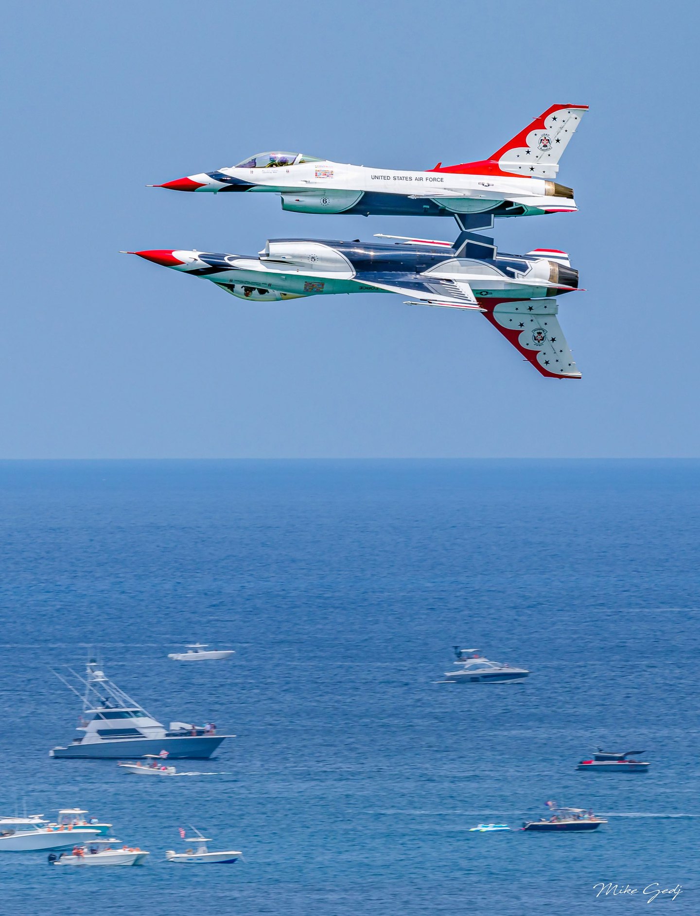 Two red, white, and blue U.S. Air Force Thunderbirds jets fly in close formation, one inverted beneath the other, above a blue sea dotted with boats.
