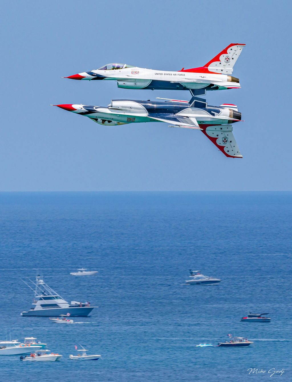 Two red, white, and blue U.S. Air Force Thunderbirds jets fly in close formation, one inverted beneath the other, above a blue sea dotted with boats.