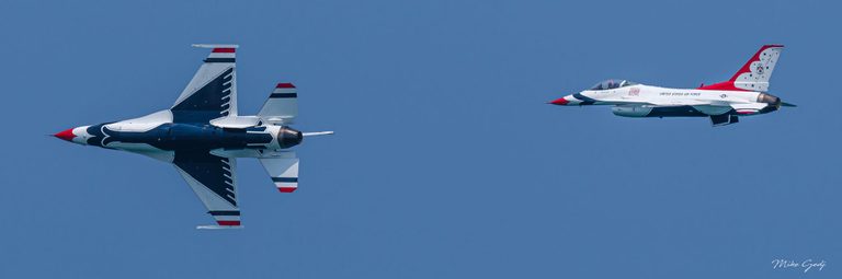 Two U.S. Air Force Thunderbirds jets fly leftward against a clear blue sky, the nearer aircraft banking steeply to reveal its patterned underside.