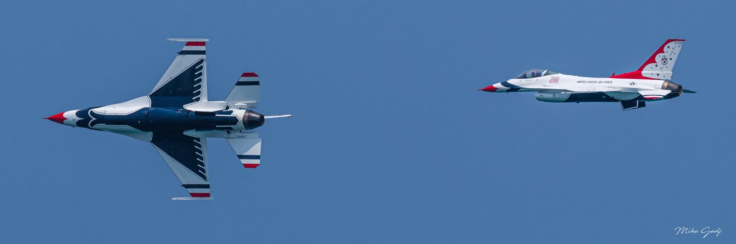 Two U.S. Air Force Thunderbirds jets fly leftward against a clear blue sky, the nearer aircraft banking steeply to reveal its patterned underside.