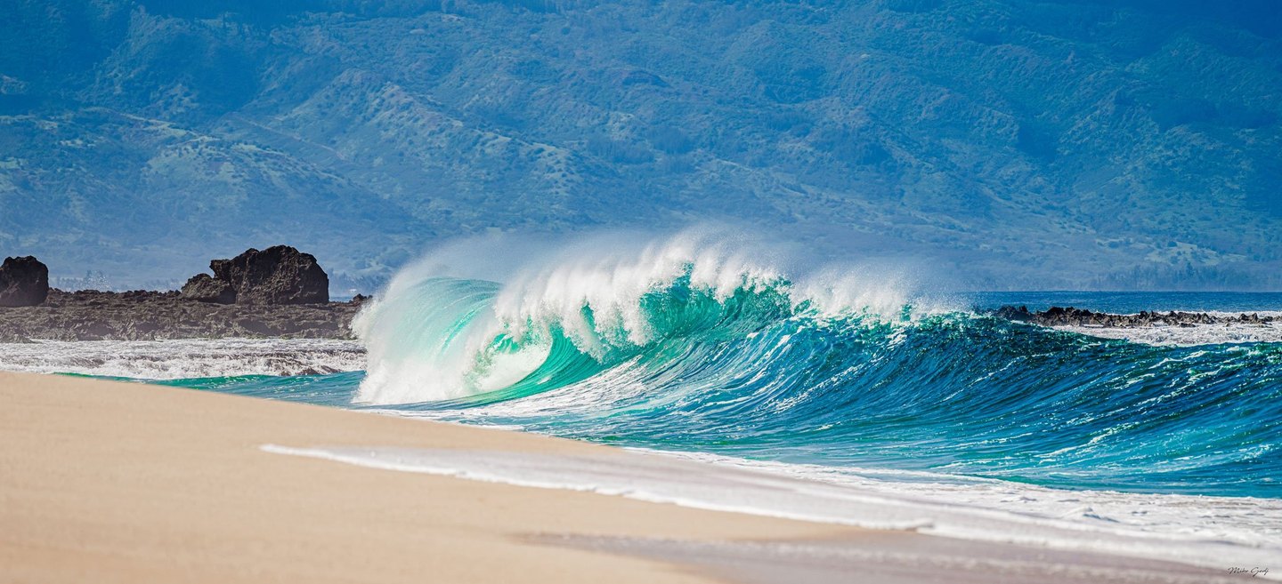 A turquoise wave breaks beside a pale sandy beach, throwing white spray above dark shoreline rocks with hazy mountain slopes in the background.