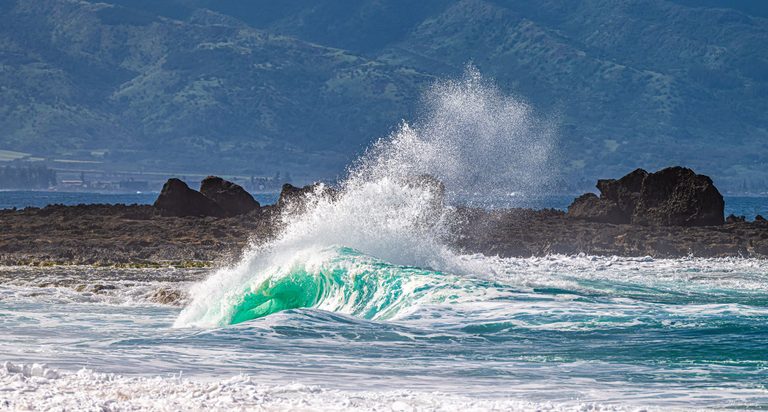 A turquoise wave curls in front of a dark rocky shoreline as white spray rises against a backdrop of green mountain slopes.
