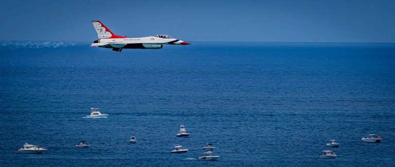 A red, white, and blue U.S. Air Force Thunderbird jet flies above a deep blue sea dotted with small motorboats.