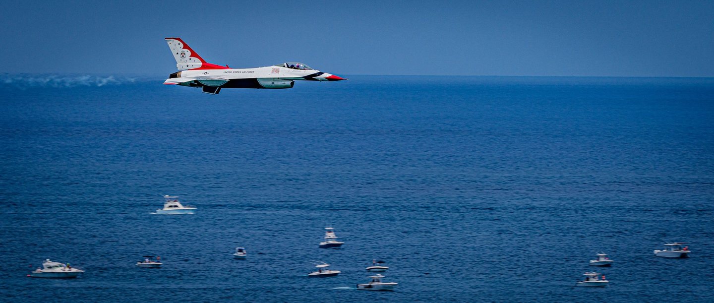 A red, white, and blue U.S. Air Force Thunderbird jet flies above a deep blue sea dotted with small motorboats.