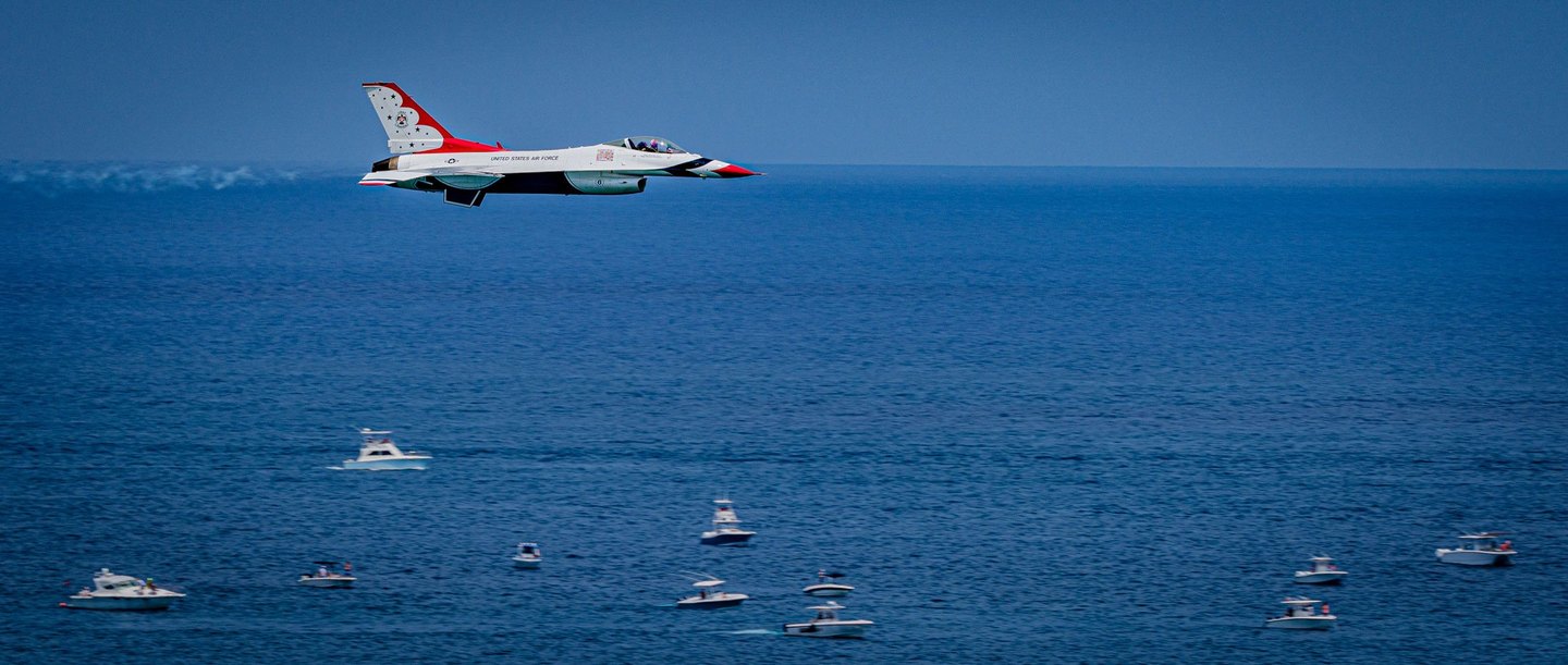 A red, white, and blue U.S. Air Force Thunderbird jet flies above a deep blue sea dotted with small motorboats.