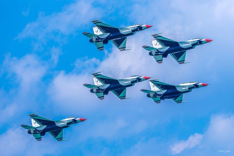 Five red, white, and blue fighter jets fly in formation toward the right, viewed from below against a bright blue sky with scattered white clouds.