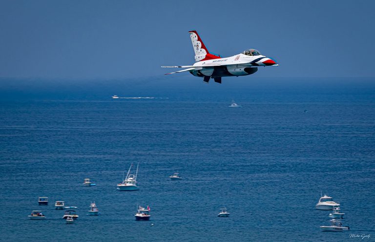 A red, white, and blue U.S. Air Force Thunderbirds F-16 flies over a blue ocean dotted with recreational boats.