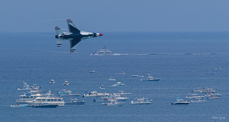 A red, white, and blue fighter jet banks over a blue sea crowded with motorboats and yachts, with faint vapor trails extending behind its wings.