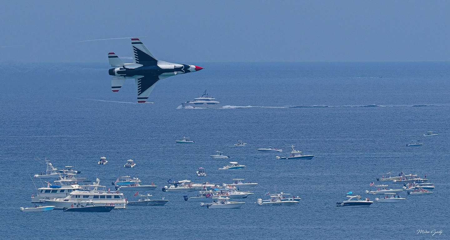 A red, white, and blue fighter jet banks over a blue sea crowded with motorboats and yachts, with faint vapor trails extending behind its wings.
