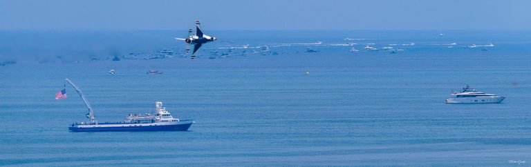 A red, white, and blue jet banks above a blue sea dotted with boats, with a large vessel flying an American flag at left and a white yacht at right.