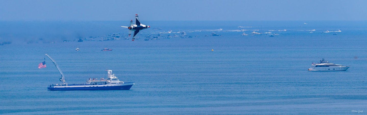 A red, white, and blue jet banks above a blue sea dotted with boats, with a large vessel flying an American flag at left and a white yacht at right.
