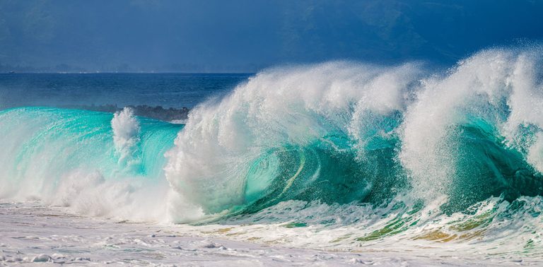 A large turquoise wave breaks into white spray above foamy surf, with deep blue water and hazy mountains in the background.