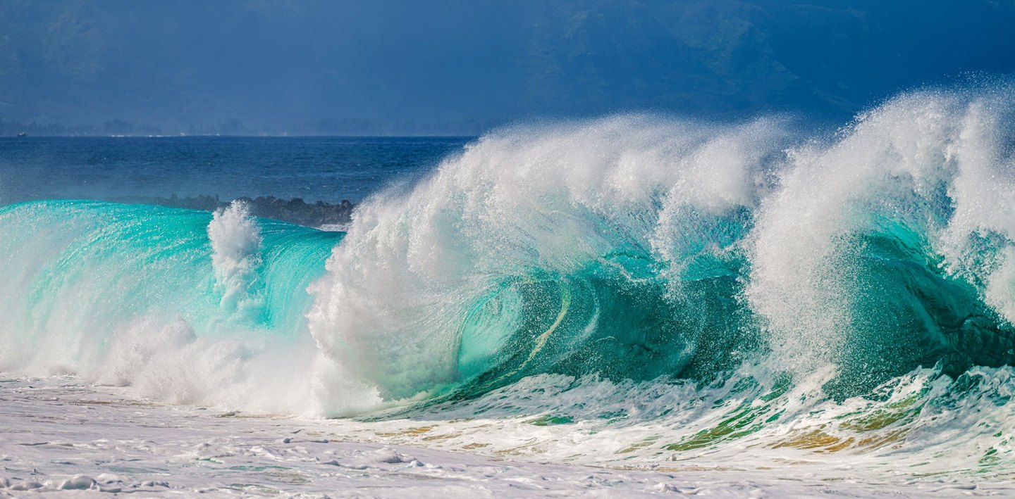 A large turquoise wave breaks into white spray above foamy surf, with deep blue water and hazy mountains in the background.
