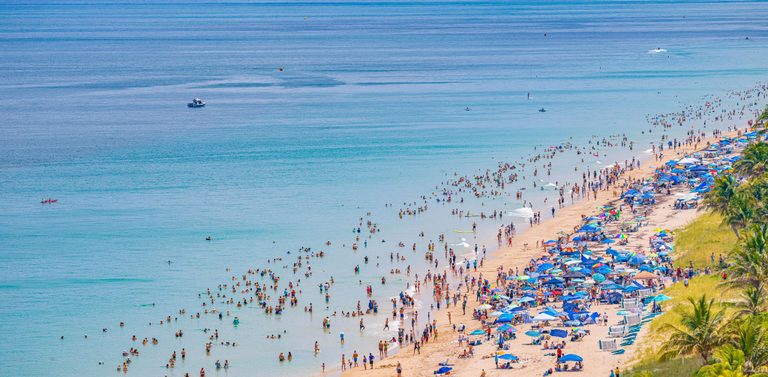 An elevated view shows a crowded sandy beach lined with colorful umbrellas and palms, with swimmers in turquoise shallows and small boats farther offshore.
