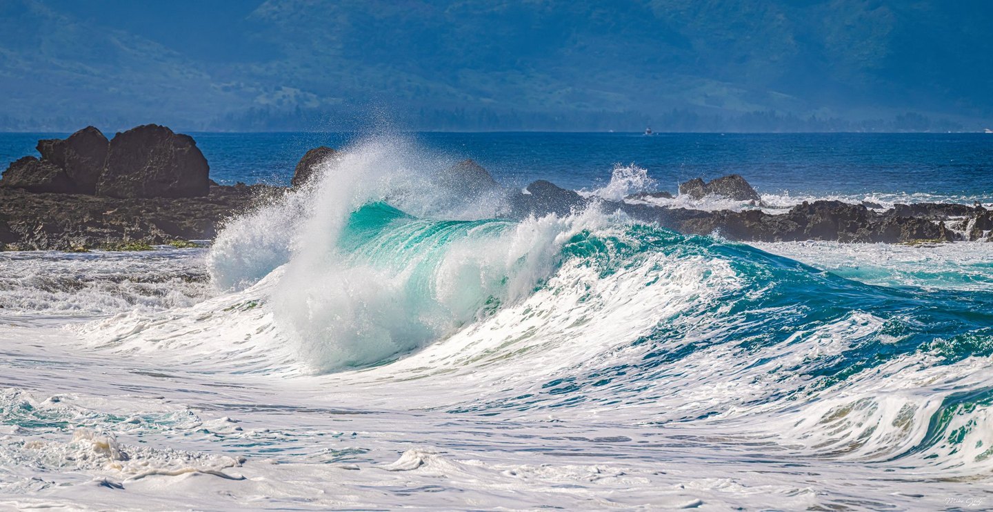 A turquoise ocean wave breaks into white spray before dark coastal rocks, with hazy mountains across the water.