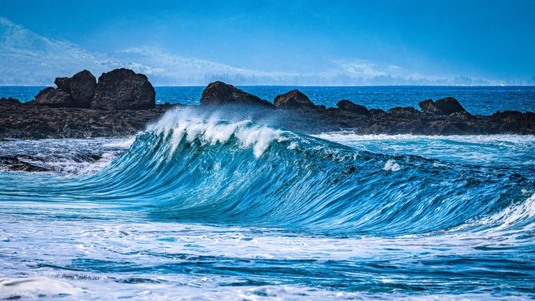 A turquoise wave crests with white spray in front of dark coastal rocks, with a hazy mountainous shoreline in the distance.