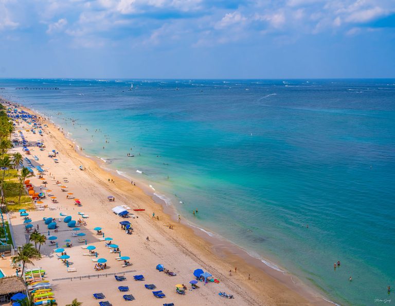 An elevated view shows a sandy beach lined with colorful umbrellas, lounge chairs, and palms beside turquoise water dotted with swimmers and distant boats.