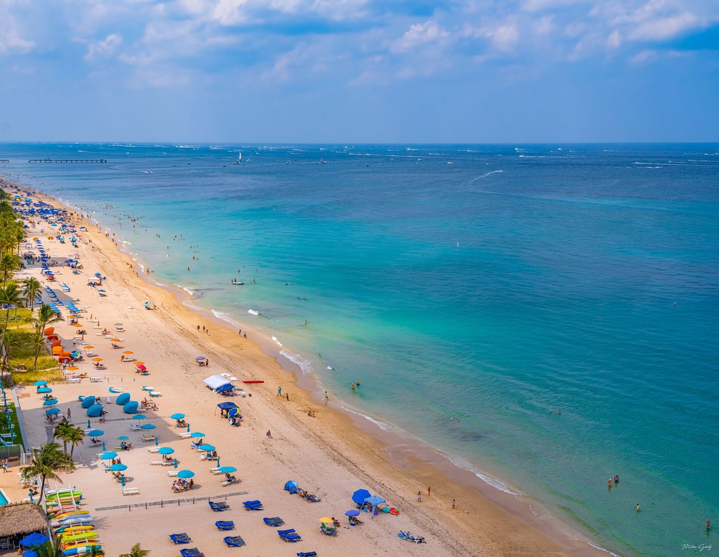 An elevated view shows a sandy beach lined with colorful umbrellas, lounge chairs, and palms beside turquoise water dotted with swimmers and distant boats.