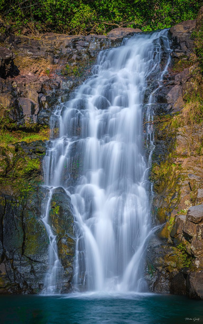 A silky white waterfall cascades over dark, moss-covered rock ledges into a turquoise pool beneath green foliage.