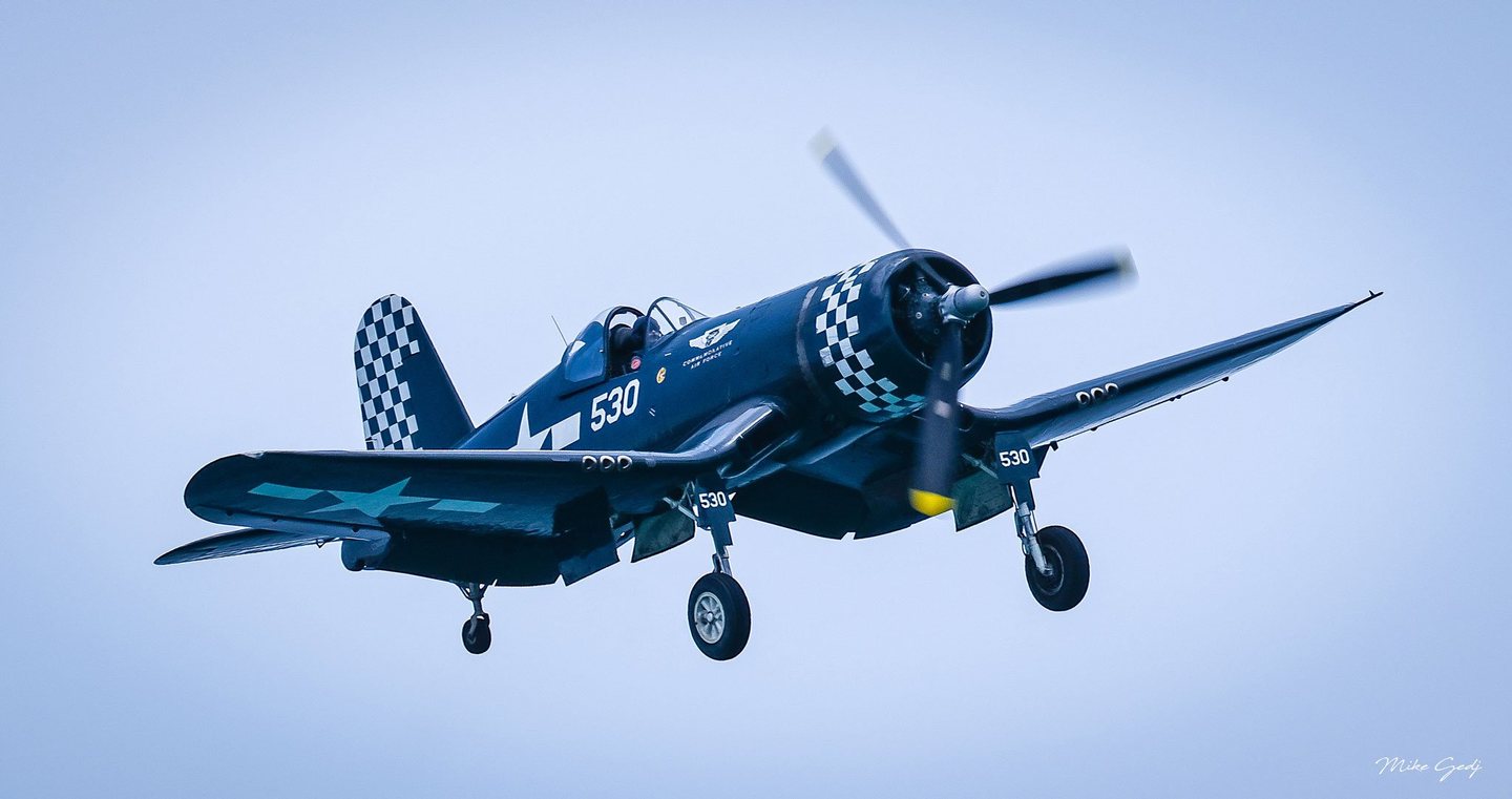 A dark blue Corsair aircraft marked 530 flies against a pale blue sky with its landing gear lowered, propeller blurred, and checkerboard markings on its nose and tail.