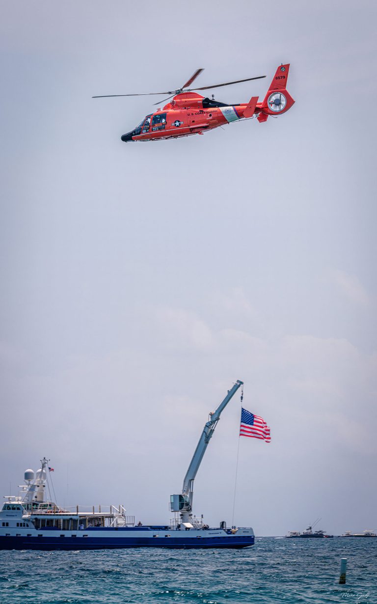 A red U.S. Coast Guard helicopter flies above a blue-and-white vessel with an American flag suspended from its crane, against a pale sky over choppy water.